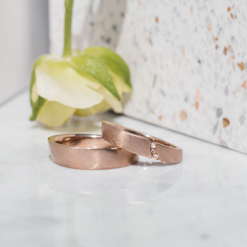 Eheringe aus 750er Roséweißgold. Der Damenring ist ein Spalt-Ring mit drei weißen Brillanten, die Oberfläche matt gestrichen.