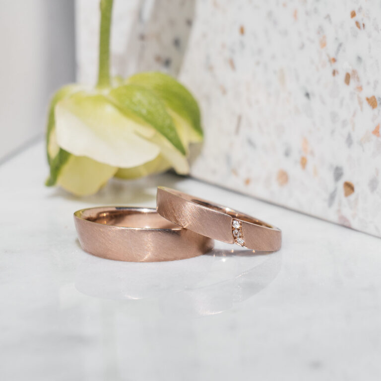 Eheringe aus 750er Roséweißgold. Der Damenring ist ein Spalt-Ring mit drei weißen Brillanten, die Oberfläche matt gestrichen.