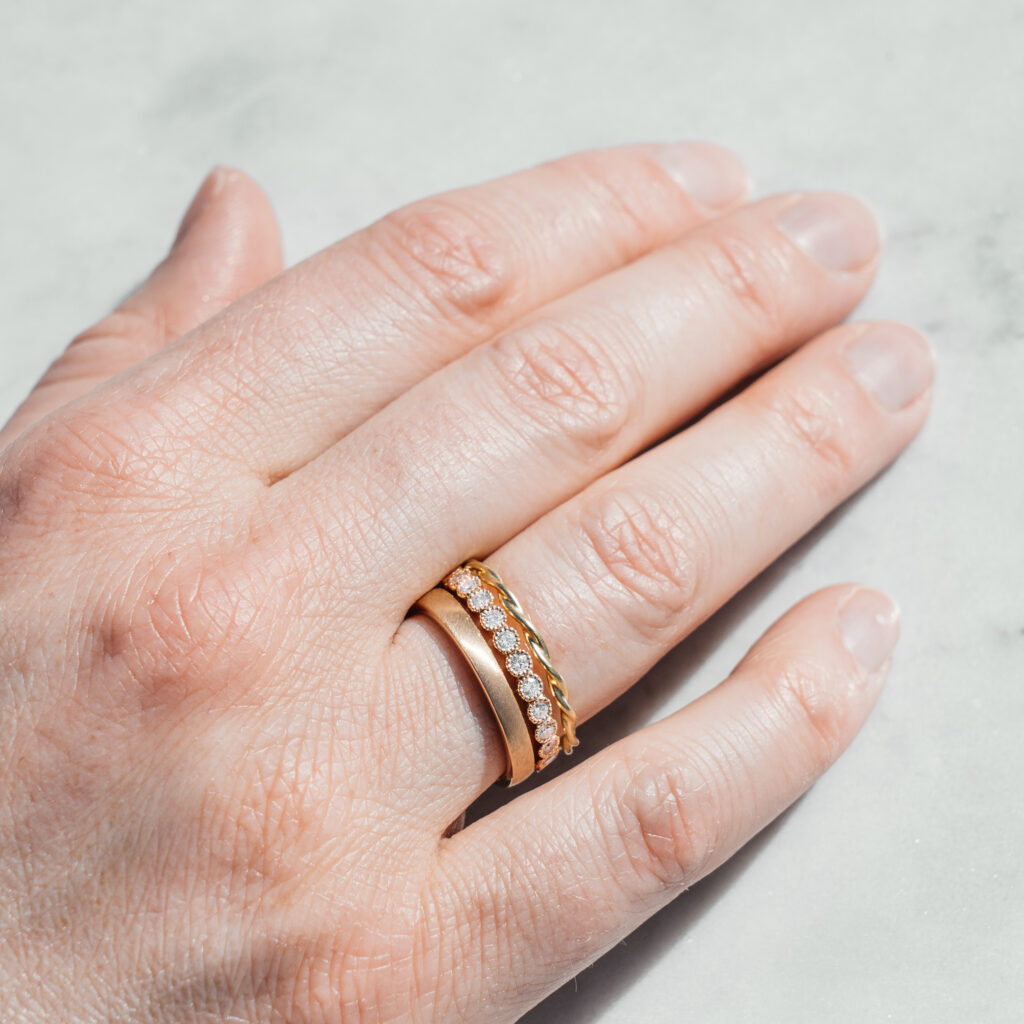 Eine Hand, die drei Ringe am Ringfinger trägt. Ein feiner goldener Kordelring, einer mit filigran gefassten Brillanten und ein schlichter schmaler Ring mit matter Oberfläche.