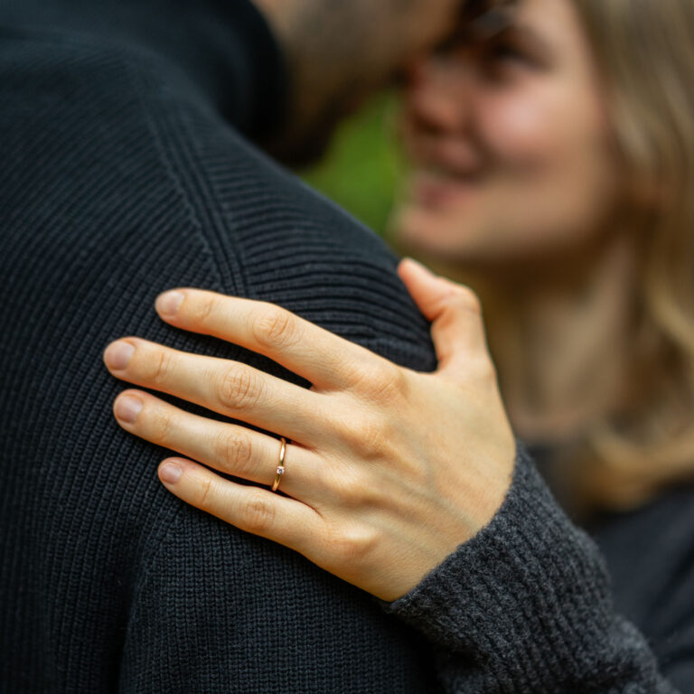 Hand einer Frau auf der Schulter des Mannes, sie trägt einen zarten Verlobungsring aus Gelbgold mit Brillant am Ringfinger.
