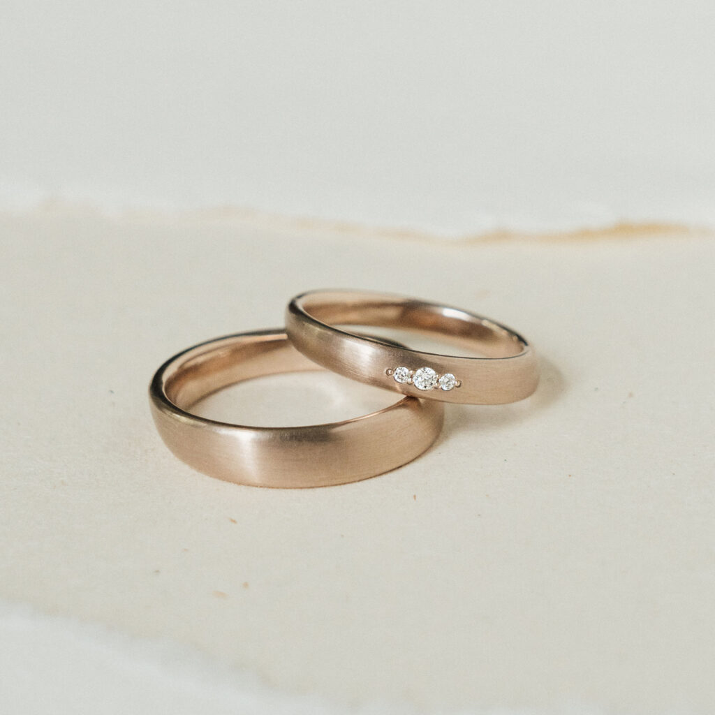 Ergonomische Eheringe aus Roséweißgold mit drei Brillanten im Damenring. Matte Oberfläche