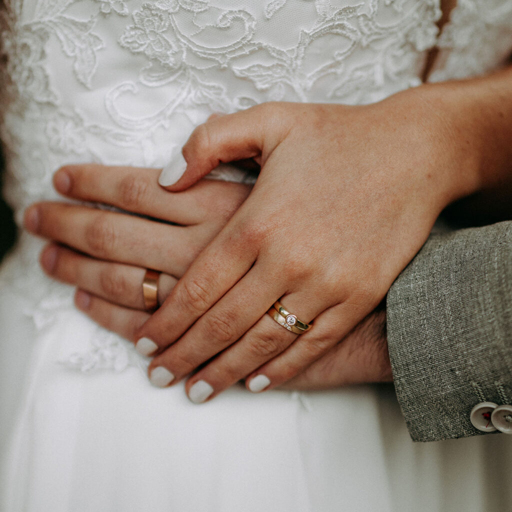 Hände eines Hochzeitspaars mit ergonomischen Eheringen. Die Braut trägt ihren Verlobungsring mit Brillant in Kombination mit ihrem Ehering.