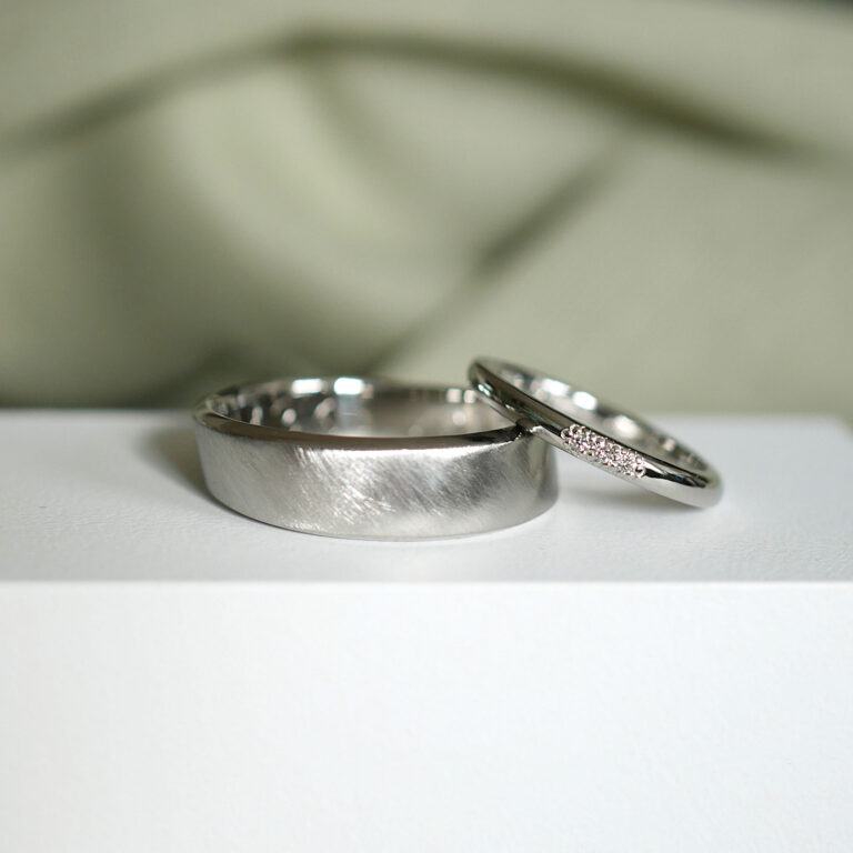Zwei ergonomische Eheringe, einer mit strichmatter Oberfläche, einer glänzend poliert mit drei Brillanten in Blümchen Fassung. Die Ringe sind aus Platin.