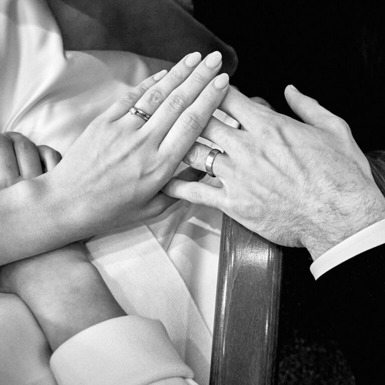 Die Hände eines Ehepaars bei der Trauung mit Eheringen. Die Braut trägt ihren Ehering in Kombination mit ihrem Verlobungsring mit Brillant.