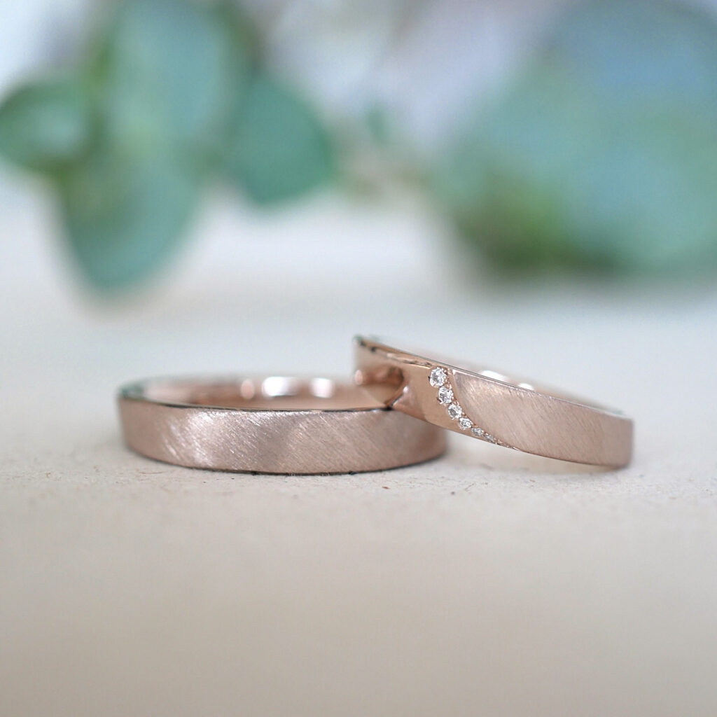 Zwei ergonomische Eheringe aus Roséweißgold mit strichmatter Oberfläche. Im Damenring eine Brillantenreihe.