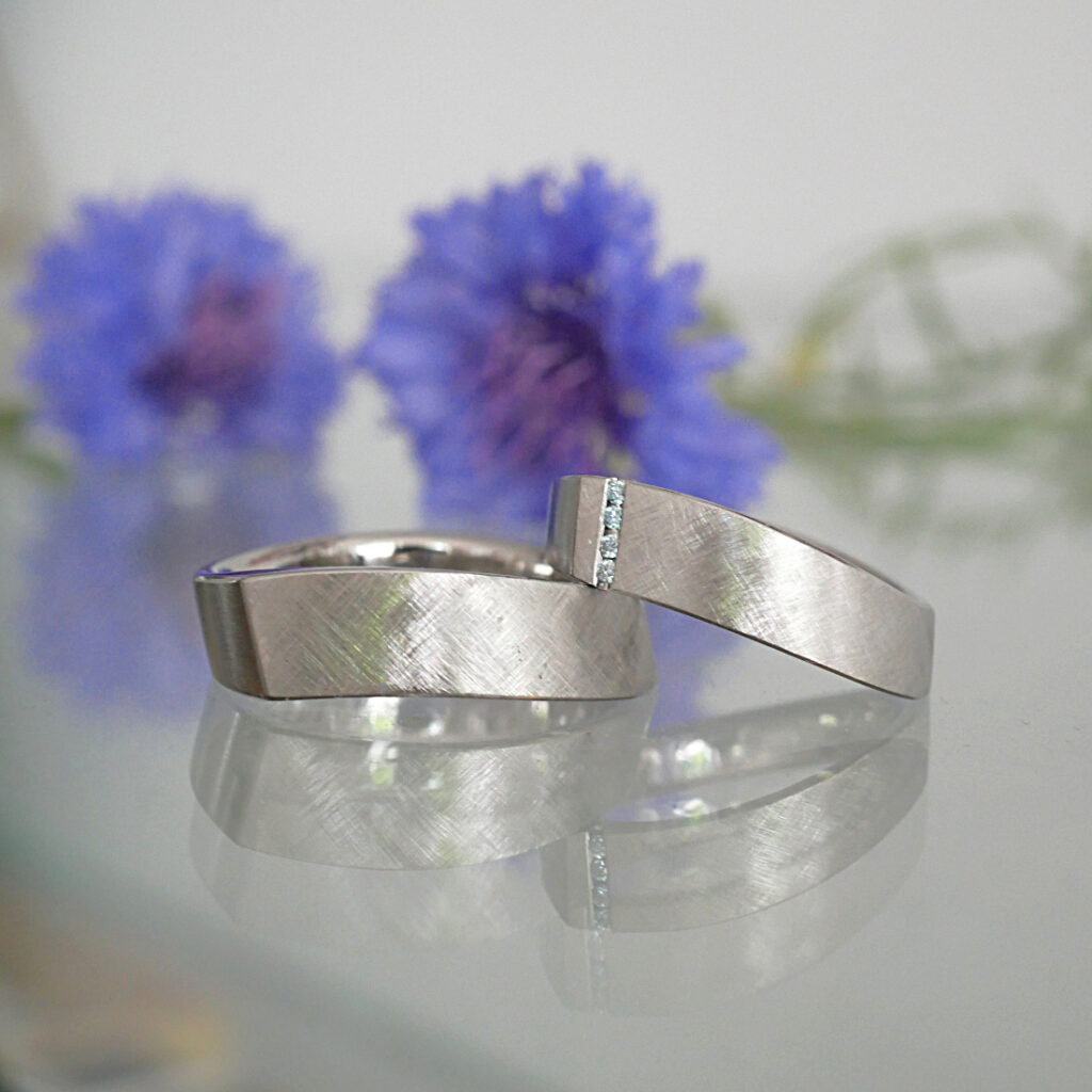 Zwei ergonomische Eheringe mit besonderer Form aus Platin. Kreuzmatte Oberfläche und Brillanten im Damenring.